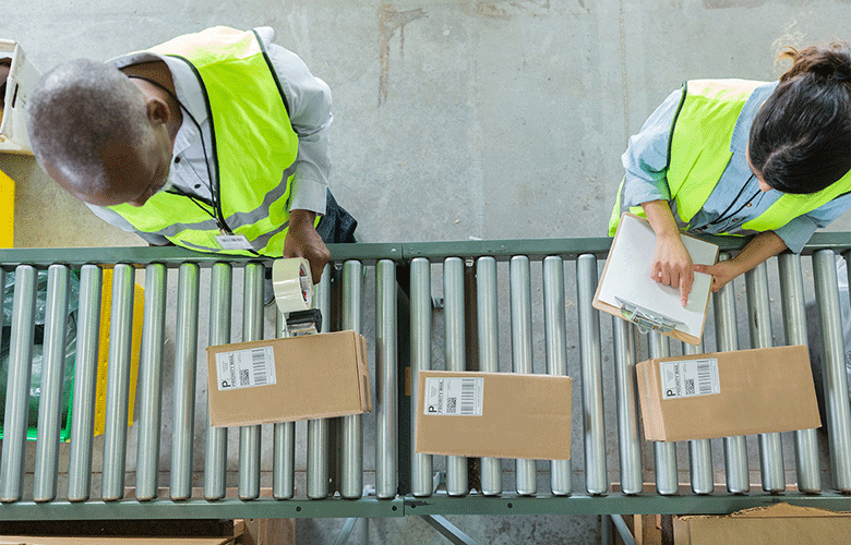 Man with tape gun standing over boxes on assembly line with woman holding clipboard in high vis vests