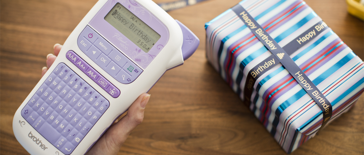 Hand holding purple p-touch labelling device with a table in background with a gift-wrapped box tied with printed ribbon