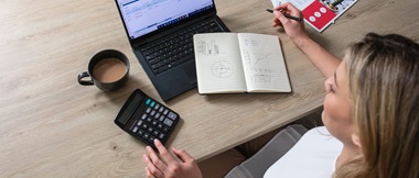 A female business owner calculating costs while sat at a desk with a notebook, calculator and laptop computer