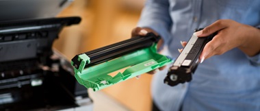 A lady holding two parts of a toner cartridge while standing next to an open laser printer