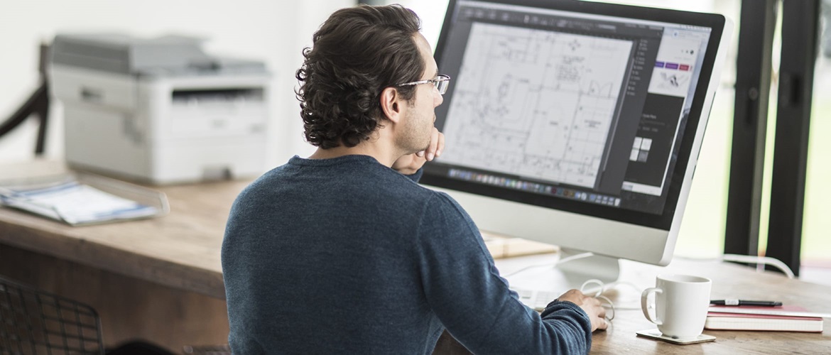 A man printing a document from an all-in-one computer while sitting at a desk in a home office environment