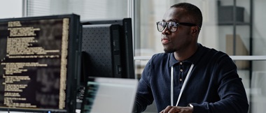 An IT decision maker working on a computer, with a second computer displaying code