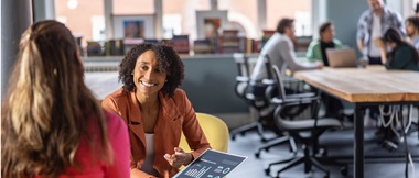 Two female colleagues discussing printed statistics in an office environment with a meeting around a desk in the background