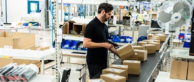 Male warehouse worker scanning a barcode adhered to a brown cardboard box as it's moving along a line on a conveyor belt