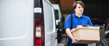 A male delivery driver wearing a blue t-shirt walking away from a white van while carrying a brown cardboard box