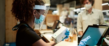 A female cashier wearing PPE while checking out products in a shop with a male customer in the background