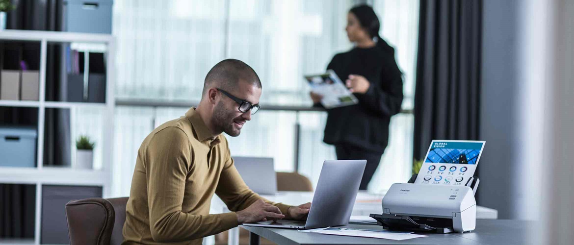 Male sat working on a laptop with a scanner and lady in the background reading