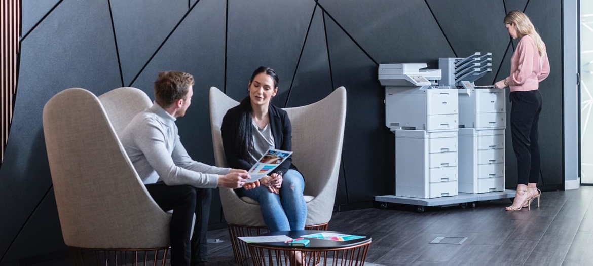 Male and female sat talking around small table, female in background stood next to printers