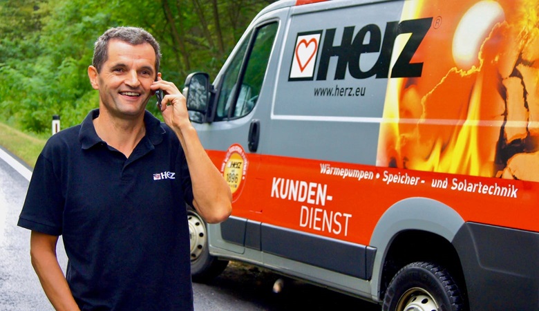 A driver for Herz talking on a mobile phone while stood next to his van, parked at the roadside with greenery in the background