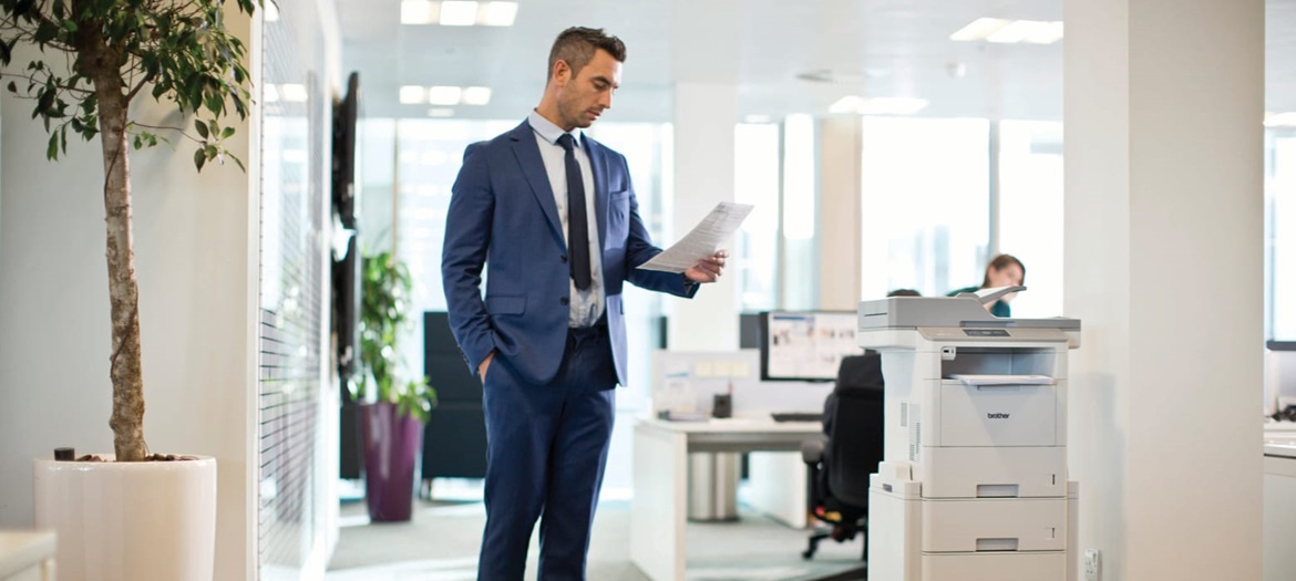 A businessman reading a document while standing next to a Brother laser printer in an office environment