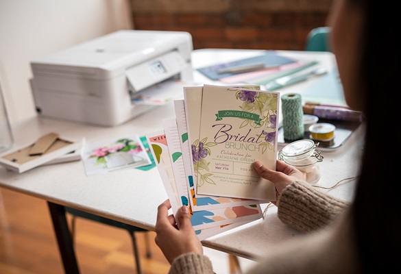 Woman holding several colourful cards with a printer and rolls of string on a table in the background