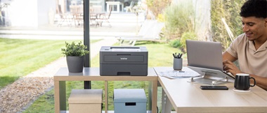 man sat at organised desk looking at laptop next to a printer and boxes on top of a sidetable with a garden in the background