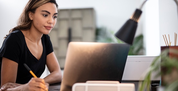 Woman holding a pen looking at a laptop with a lamp behind it