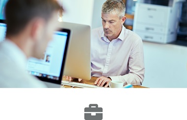 Two men sat at desk working on computers with laser printer in the background